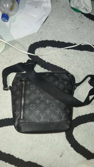 Svart axelväska  - Snygg svart axelväska med klassiskt monogrammönster och justerbar axelrem. Väskan har en dragkedja framtill och är tillverkad i skinn med detaljer i syntet. Perfekt för dig som vill ha en stilren och trendig accessoar.