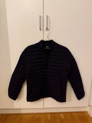 Mörkblå dunjacka från UBR - Snygg mörkblå dunjacka från UBR med minimalistisk design. Jackan har dragkedja framtill, två sidofickor med dragkedja och en hög krage med tryckknapp. Perfekt för kalla vinterdagar och enkel att matcha med olika outfits.