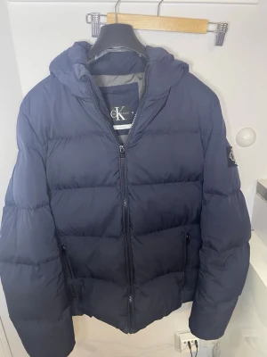  dunjacka Calvin Klein  - Snygg mörkblå dunjacka från Calvin Klein Jeans med huva och dragkedja framtill. Jackan har två sidofickor med dragkedja och en patch med logga på ärmen. Fylld med dun och fjäder för extra värme under kalla dagar.