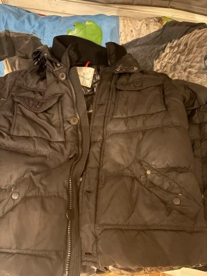 Svart dunjacka  - Snygg svart dunjacka med flera fickor framtill, hög krage och dragkedja. Jackan har en klassisk puffer-modell och på insidan. Perfekt för kalla vinterdagar och riktigt varm. PRIS KAN DISKUTERAS 