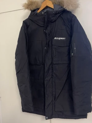 Svart parkas från Everest med fuskpäls - Säljer en svart parkas från Everest med avtagbar huva och fuskpäls. Jackan har flera stora fickor framtill, dragkedja och knappar, samt en ficka med dragkedja på ärmen. Perfekt för kalla dagar och riktigt snygg streetstil.