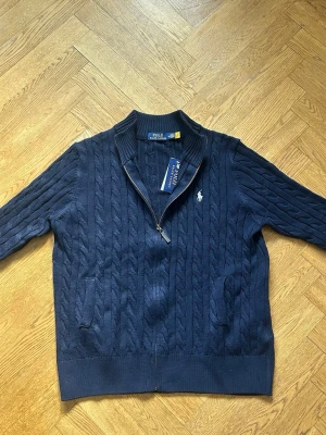 Marinblå kabelstickad kofta Polo Ralph Lauren. PASSAR M/L - Kabelstickad marinblå kofta från Polo Ralph Lauren med dragkedja framtill, två fickor och vit broderad logga på bröstet. Klassisk ribbad mudd vid ärmslut och nederkant. Perfekt för dig som gillar stilren och tidlös design. Passar M/L