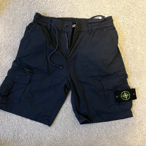 Blåa cargo shorts från Stone Island - Blåa cargo shorts från Stone Island med ikonisk patch på benet. De har flera fickor, snörning i midjan och dragkedja fram. Materialet är 97% bomull och 3% spandex, vilket gör dem både bekväma och slitstarka. Perfekta för dig som gillar streetwear och funktionella detaljer. en ficka sönder