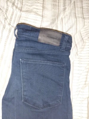 Mörkblå jeans från J.Lindeberg - Säljer ett par mörkblå jeans från J.Lindeberg med klassisk femficksdesign och diskret JL-logga vid fickan. Jeansen har normal midja och raka ben. Materialet känns mjukt och stretchigt, perfekt för en avslappnad men snygg look. W31 L32