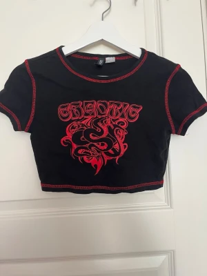 Svart croppad topp med röd drakprint - Svart croppad topp med röda kontrastsömmar och tryck framtill med texten 'CHAOTIC' och en röd orm med flera huvuden. T-shirten har korta ärmar och en edgy vibe, perfekt för dig som gillar statement-plagg.