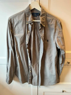 Grå skjorta från Ralph Lauren - Klassisk grå skjorta från Ralph Lauren med slim fit-passform. Skjortan har långa ärmar, knappar framtill och den ikoniska broderade loggan på bröstet. Perfekt till både jeans och chinos för en clean och stilren look. Den har lite små fläckar på ena armen som man ser på bild 5