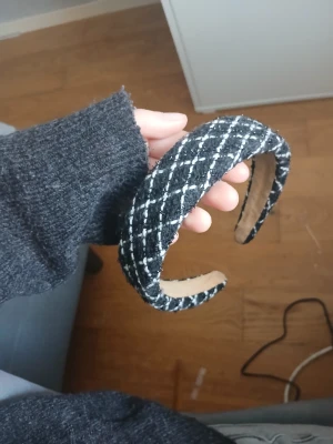 Svartvitt rutigt diadem i tweed - Snyggt svart diadem med vitt rutmönster i tweedliknande tyg. Diademet är vadderat och har en bred form som ger en trendig look. Insidan är beige och mjuk för extra komfort. Perfekt accessoar för att lyfta din outfit.