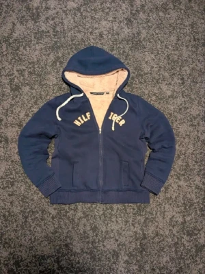 Blå hoodie från Tommy Hilfiger - Mysig marinblå hoodie från Tommy Hilfiger med dragkedja och vit text på bröstet. Insidan är fodrad med mjuk beige fleece för extra värme. Hoodien har långa ärmar, ribbade muddar och vita snören i huvan. Perfekt för kalla dagar! Storlek L men passar som S/M. 55 cm lång och 60 cm genom bröstet