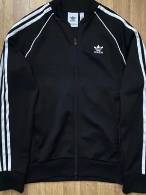 Svart Adidas track jacket - Klassisk svart track jacket från Adidas med vita ränder längs ärmarna och dragkedja framtill. Tröjan har ståkrage, ribbade muddar och Adidas logga broderad på bröstet. Perfekt för en sportig och avslappnad stil.