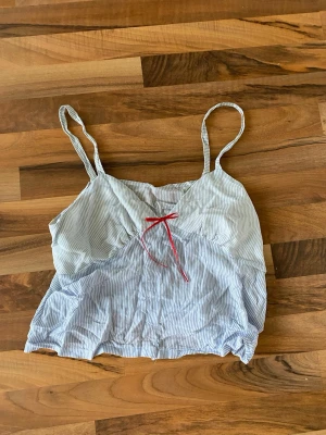 Randig linnetopp med röd rosett - Söt linnetopp med tunna axelband och smala blå och vita ränder. Toppen har en liten röd rosett framtill som detalj och är tillverkad i ett lätt och luftigt material, perfekt för varma dagar.