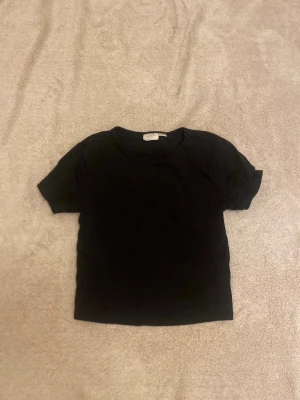 Svart basic t-shirt från Gina Tricot - En enkel svart t-shirt från Gina Tricot i storlek 158/164. T-shirten har rund halsringning och korta ärmar. Perfekt att matcha med jeans eller kjol för en clean och stilren look.