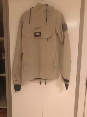 Beige vindjacka från Dope - Snygg beige vindjacka från Dope med huva och svarta detaljer. Stor ficka framtill med Dope-logga, patch på ärmen och justerbar nederkant. Jackan har muddar vid ärmsluten och dragkedjor på sidorna. Perfekt för blåsiga dagar.