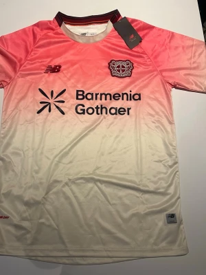 Bayer Leverkusen tröja rosa/vit New Balance - Snygg Bayer Leverkusen matchtröja från New Balance i rosa och vitt med gradient-effekt. Tröjan har klubbmärke och sponsortryck på bröstet samt korta ärmar. Tillverkad i lätt och ventilerande funktionsmaterial, perfekt för träning eller match.