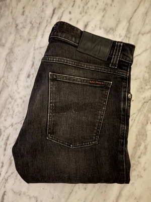 Nudie jeans - Snygga mörkgråa jeans från Nudie Jeans med diskret tvättad look och klassisk femficksdesign. Jeansen har detaljerade sömmar, samt en svart läderpatch bak i midjan. Perfekta för en stilren och avslappnad outfit.