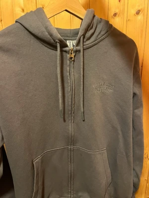 Grå zip hoodie - Mörkgrå hoodie med dragkedja. Modellen har huva med dragsko, två stora fickor framtill och broderad text 'ATHLETIC CLUB' på bröstet. Perfekt för en avslappnad och sportig stil.