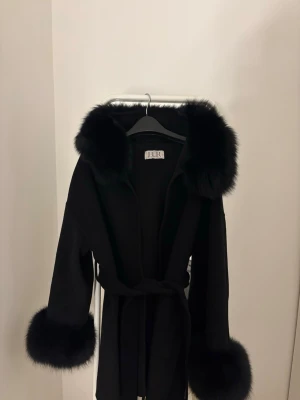 Furstockholm jacka - Säljer en svart pälsjacka från FUR med stora fluffiga pälskanter vid huva och ärmslut. Jackan har knytband i midjan för snygg passform. Perfekt för dig som vill ha en statement jacka till vintern.