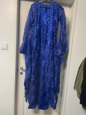 Kurdisk klänning  - En blå långklänning med glittriga detaljer och ett snyggt mönster. Klänningen är ärmlös med en matchande transparent överdel med långa ärmar. Perfekt för dig som vill sticka ut med en elegant look.
