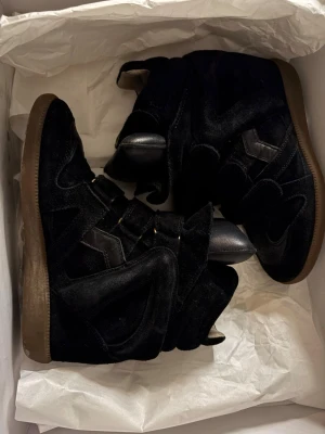 Svarta Isabel Marant wedge sneakers - Snygga svarta sneakers från Isabel Marant med högt skaft och kilklack. Skorna är gjorda i mocka och läder med coola paneldetaljer och kardborreband. Perfekta för dig som vill ha en trendig och unik look.