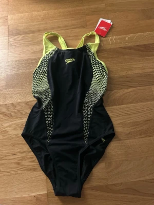 Svart och gul baddräkt från Speedo - Sportig baddräkt från Speedo i svart med gula detaljer och mönstrade sidor. Axelbanden är gula och modellen har en klassisk, sportig passform som passar perfekt för simträning. Materialet känns som syntet och baddräkten har ett coolt, grafiskt mönster på sidorna.