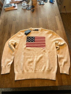 Vit tröja med USA-flagga Ralph Lauren - Vit stickad tröja från Ralph Lauren med stor amerikansk flagga på bröstet och RL-broderi. Tröjan har rund halsringning, ribbade muddar och nederkant. Perfekt för dig som gillar klassisk och stilren design med en amerikansk touch.