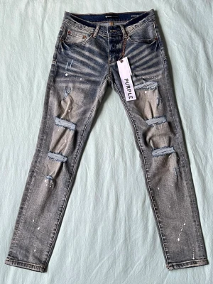 Grå slitna jeans från Purple Brand - Säljer ett par grå skinny jeans från Purple Brand med slitningar och hål på benen samt vita färgstänk. Jeansen har fem fickor, dragkedja och knappgylf. Snygg tvätt med tydliga kontraster och trendig streetkänsla.