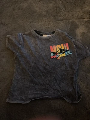 Grå t-shirt från Zara med tryck - Grå t-shirt från Zara med färgglatt tryck 'Maui and Sons' på bröstet. T-shirten har en tvättad look och klassisk rund hals. Perfekt för en avslappnad stil och enkel att matcha med jeans eller shorts.