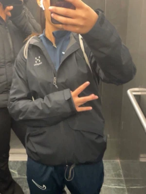 Svart vindjacka från Haglöfs - Svart vindjacka från Haglöfs med dragkedja framtill och vit logga på bröstet. Jackan har hög krage, normal passform och är gjord i lätt syntetmaterial. Perfekt för blåsiga dagar och enkel att ta med sig.