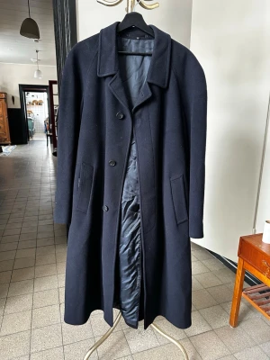 Mörkblå Kashmir kappa - Stilren mörkblå kashmirkappa med klassisk krage och tre knappar framtill. Kappan har två snedställda fickor och ett avtagbart bälte i midjan för en snygg siluett. Fodrad insida för extra komfort. Perfekt för dig som vill ha en tidlös och elegant look.