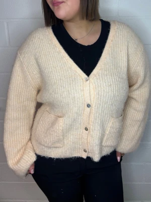 Ljusbeige fluffig kofta med fickor - Mysig ljusbeige kofta i ribbstickat material med fluffig känsla. Koftan har v-ringning, knäppning framtill med silverfärgade knappar och två praktiska fickor. Perfekt för lager på lager och en avslappnad look.