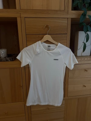 Vit träningstopp från Stronger - Snygg vit träningstopp från Stronger med korta ärmar och rund halsringning. Tillverkad i ett stretchigt syntetmaterial som andas, perfekt för aktiva dagar. Diskret logga på bröstet och figurnära passform.