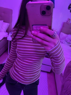 Randig långärmad topp med knappar - Säljer en beige och vit randig långärmad topp med dekorativa knappar på axeln. Toppen har en slim passform och är tillverkad i ett mjukt, ribbat material som sitter snyggt på kroppen. Perfekt att styla med jeans eller kjol.