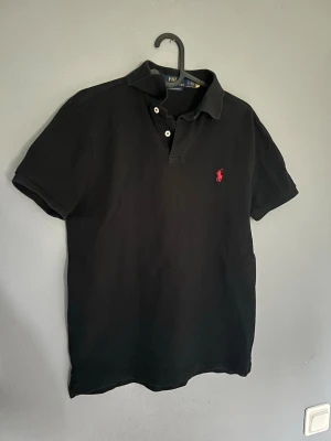 Svart pikétröja Polo Ralph Lauren - Klassisk svart pikétröja från Polo Ralph Lauren med röd broderad logga på bröstet. Passar riktigt bra till sommaren🤩 skicket är väldigt bra och har inga slitningar. Storlek S. Hör av er för fler frågor😃