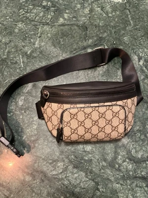 Beige axelväska från Gucci - Snygg beige axelväska från Gucci med klassiskt GG-mönster och svarta detaljer. Väskan har justerbart band, dragkedja upptill och en praktisk framficka. Perfekt för dig som vill ha både stil och funktion. Materialet är canvas med läderdetaljer. För fler bilder eller frågor kom dm:)