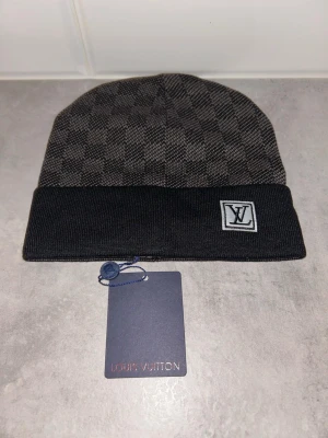 Svart och grå Louis Vuitton mössa - Snygg svart och grå mössa från Louis Vuitton med klassiskt rutmönster och vikt kant. Mössan har en LV-logga framtill och är tillverkad i mjukt, stickat material som känns lyxigt och stilrent.