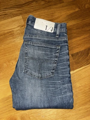 Blå slim fit jeans från T/J - Snygga blå slim fit jeans från T/J med klassisk femficksdesign och diskreta slitningar. Jeansen har en något tvättad look och raka ben. Tillverkade i mjukt denimtyg som ger en bekväm passform. Perfekta för en avslappnad och trendig stil.