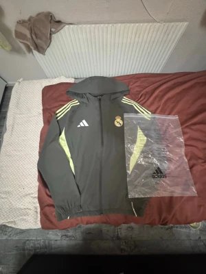 Adidas Real Madrid träningsjacka grå - Säljer en grå träningsjacka från Adidas med Real Madrid-logga på bröstet och gula detaljer längs ärmarna. Jackan har huva, dragkedja framtill och klassiska Adidas-ränder på axlarna. Perfekt för träning eller sportiga dagar.