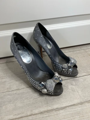 Grå pumps med strass från Karen Millen - Snygga grå pumps i storlek 36 men passar 37 från Karen Millen med öppen tå, dekorerade med glittrande strass framtill. Skorna har smal klack och är täckta av små stenar för extra bling. Perfekta för dig som vill sticka ut med elegans.
