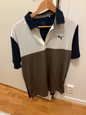 Puma golf-pikétröja i blockfärger - Snygg pikétröja från Puma med breda block i vitt, olivgrönt och mörkblått. Klassisk krage och knappslå framtill. Puma-logga broderad på bröstet. Perfekt för en sportig och avslappnad stil.