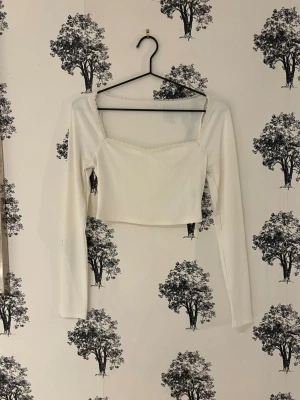 Vit croppad långärmad topp - Trendig vit croppad topp med långa ärmar och fyrkantig urringning. Toppen är ribbad och har en enkel, stilren design som passar perfekt till jeans eller kjol. Materialet känns mjukt och stretchigt för en bekväm passform.