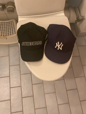 Svart och marinblå keps duo - Två snygga kepsar: en svart keps från STEINBERGS med broderad text och en klassisk marinblå New York Yankees-keps med vit logga framtill. Båda har böjd skärm och justerbar passform. Perfekta för dig som vill ha streetstyle-vibe.