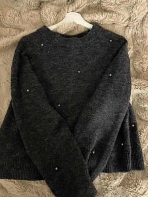 Mörkgrå stickad tröja med pärlor - Mysig mörkgrå stickad tröja med rund halsringning och små pärldetaljer utspridda över hela tröjan. Tröjan har långa ärmar och ribbstickade muddar vid ärmslut och nederkant. Perfekt för kalla dagar och enkel att matcha.