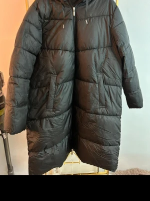 Svart lång dunjacka med huva - Säljer en svart lång dunjacka med huva och dragkedja framtill. Jackan har två snedställda fickor och quiltad design som ger en schysst look. Perfekt för kalla vinterdagar och riktigt varm tack vare dunfyllningen.