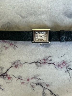 Klassisk fyrkantig klocka med svart läderband - Stilren klocka med fyrkantig silverfärgad boett och vit urtavla med romerska siffror. Klockan har ett svart läderband med struktur och klassisk design. Perfekt accessoar för en tidlös look.