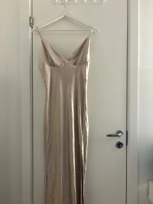 Beige långklänning från Boohoo - Säljer en elegant beige långklänning från Boohoo med smala axelband och v-ringad urringning. Klänningen har en slät, glansig yta och figurnära passform som ger en stilren siluett. Perfekt för dig som gillar minimalistisk och chic stil.