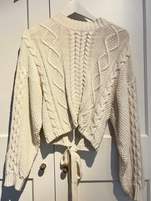 Krämvit stickad tröja med knytband - Trendig krämvit stickad tröja med grova flätmönster och ribbad halsringning. Tröjan har långa ärmar och unika knytband i midjan som ger en cool twist. Materialet känns mjukt och tjockt, perfekt för lager på lager. Passar dig som gillar en avslappnad men ändå stylish look.
