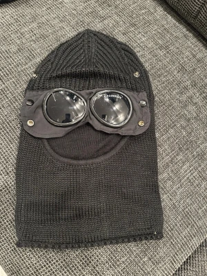 CP company  - Säljer en svart stickad balaclava med ribbad topp och slätstickad nederdel. Utrustad med detaljer i form av knappar och tygpanel på sidorna. Perfekt för kalla dagar och ger en cool look.