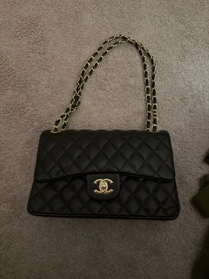  svart quiltad handväska - Säljer en klassisk svart quiltad handväska med guldfärgade detaljer och kedjerem. Väskan har det ikoniska CC-spännet framtill och röd insida med präglad logga. Tillverkad i läder och har elegant, tidlös design.