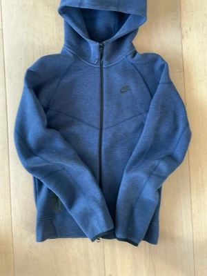 Nike tech - Mörkblå hoodie från Nike med dragkedja framtill och huva. Tillverkad i mjukt material med diskret Nike-logga på bröstet. Hoodien har långa ärmar och två sidofickor med dragkedja. Perfekt för en avslappnad och sportig stil.