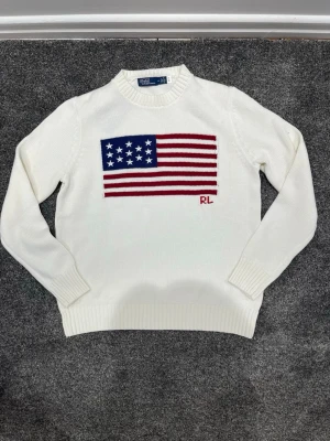 Vit stickad tröja Polo Ralph Lauren - Säljer en vit stickad tröja från Polo Ralph Lauren med amerikansk flagga i rött, vitt och blått på bröstet samt RL-broderi. Tröjan har rund halsringning och ribbade muddar vid ärmslut och nederkant. Perfekt för dig som gillar klassisk och stilren design.