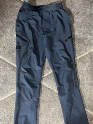 Blå träningsbyxor från Under Armour - Snygga blå träningsbyxor från Under Armour med svarta detaljer och dragkedjefickor på sidorna. Tillverkade i ett stretchigt funktionsmaterial som passar perfekt för träning eller löpning. Logga på vänster lår och modern passform.